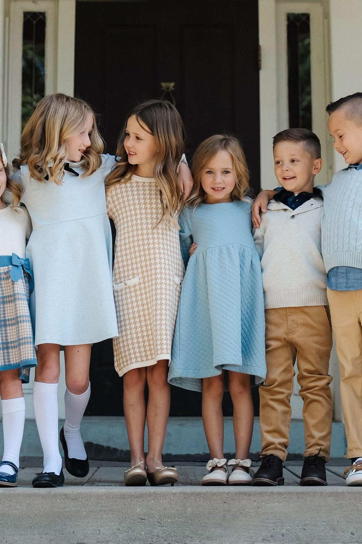A group of six children stands together, wearing stylish outfits in soft colors, including dusty blue and cream.