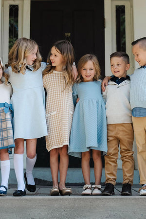 A group of six children stands together, wearing stylish outfits in soft colors, including dusty blue and cream.