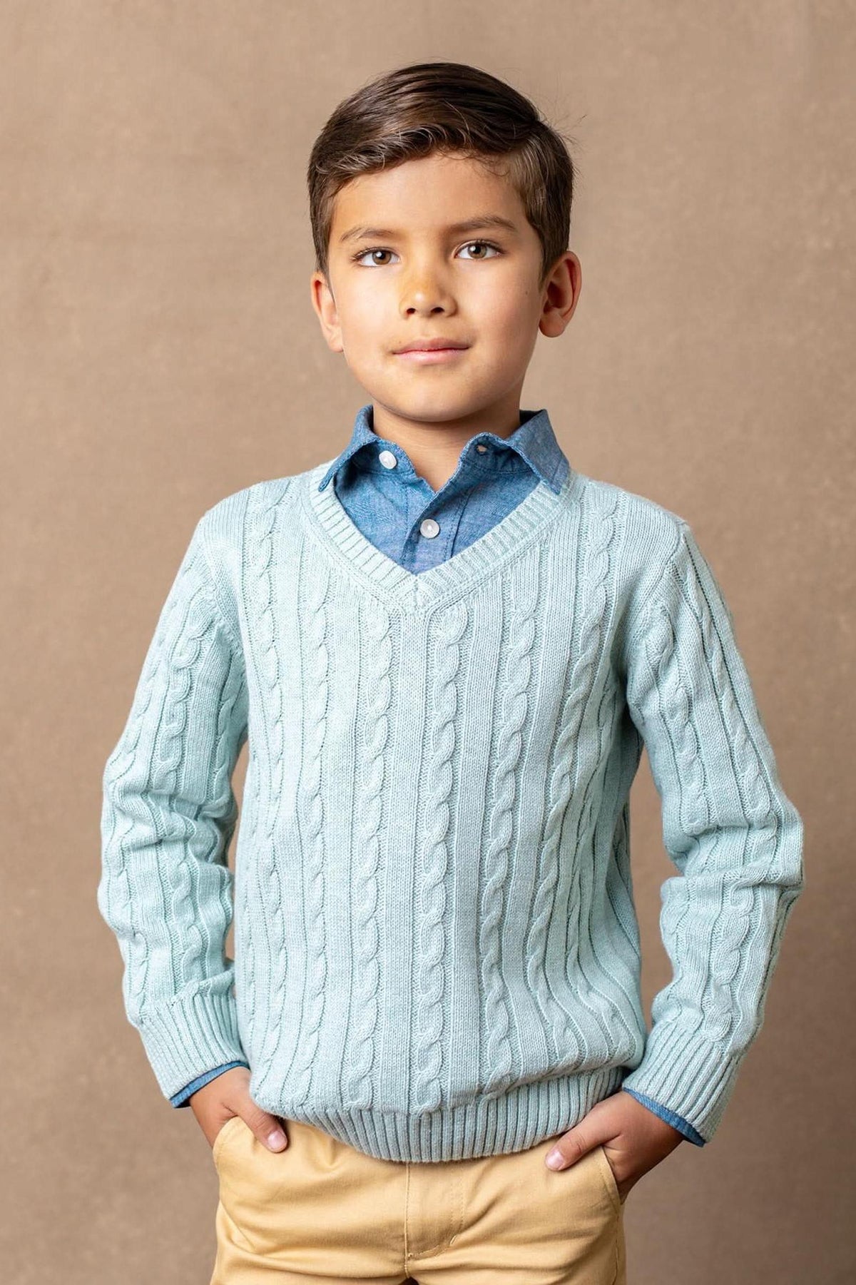 A boy wears a dusty blue organic cotton cable knit V-neck sweater, paired with a denim shirt underneath. The soft, timeless design is perfect for various occasions.