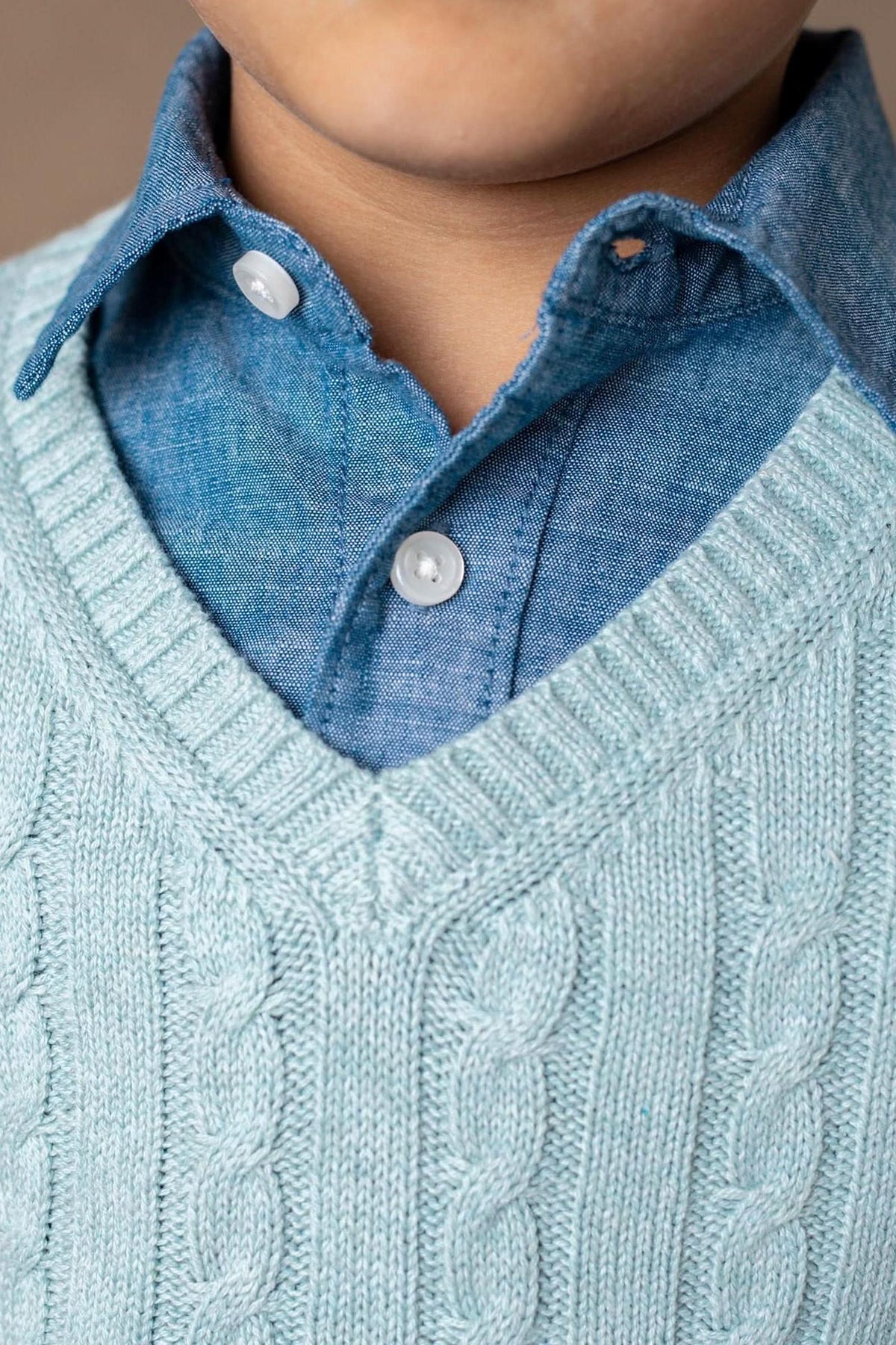 A close-up of a child wearing a dusty blue cable knit V-neck sweater over a light blue button-up shirt. The soft organic cotton fabric highlights a stylish and comfortable look.