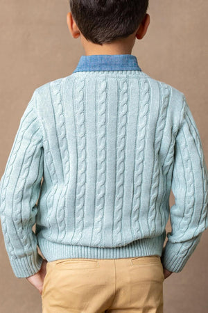 A young boy is shown from the back, wearing a dusty blue cable knit sweater made from soft organic cotton, paired with a collared shirt.
