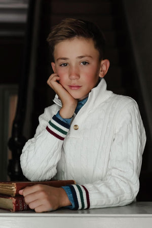 A young boy poses thoughtfully, resting his chin on his hand while wearing a white cable-knit sweater.