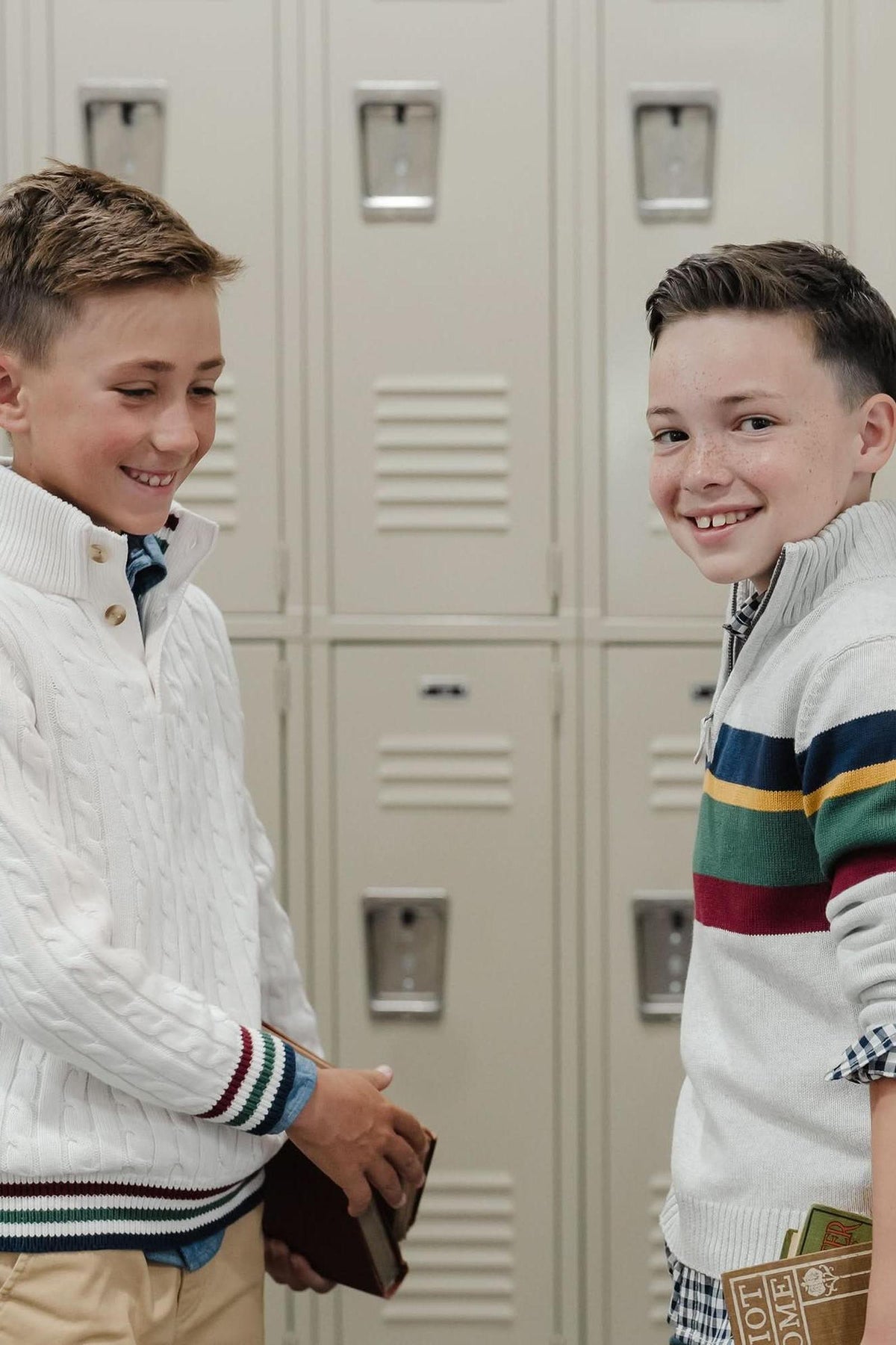Two boys are smiling and standing in front of beige school lockers. One boy wears a white cable-knit sweater, while the other sports a striped mock neck sweater, both suitable.