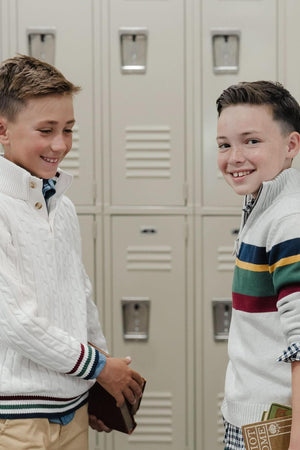 Two boys are smiling and standing in front of beige school lockers. One boy wears a white cable-knit sweater, while the other sports a striped mock neck sweater, both suitable.