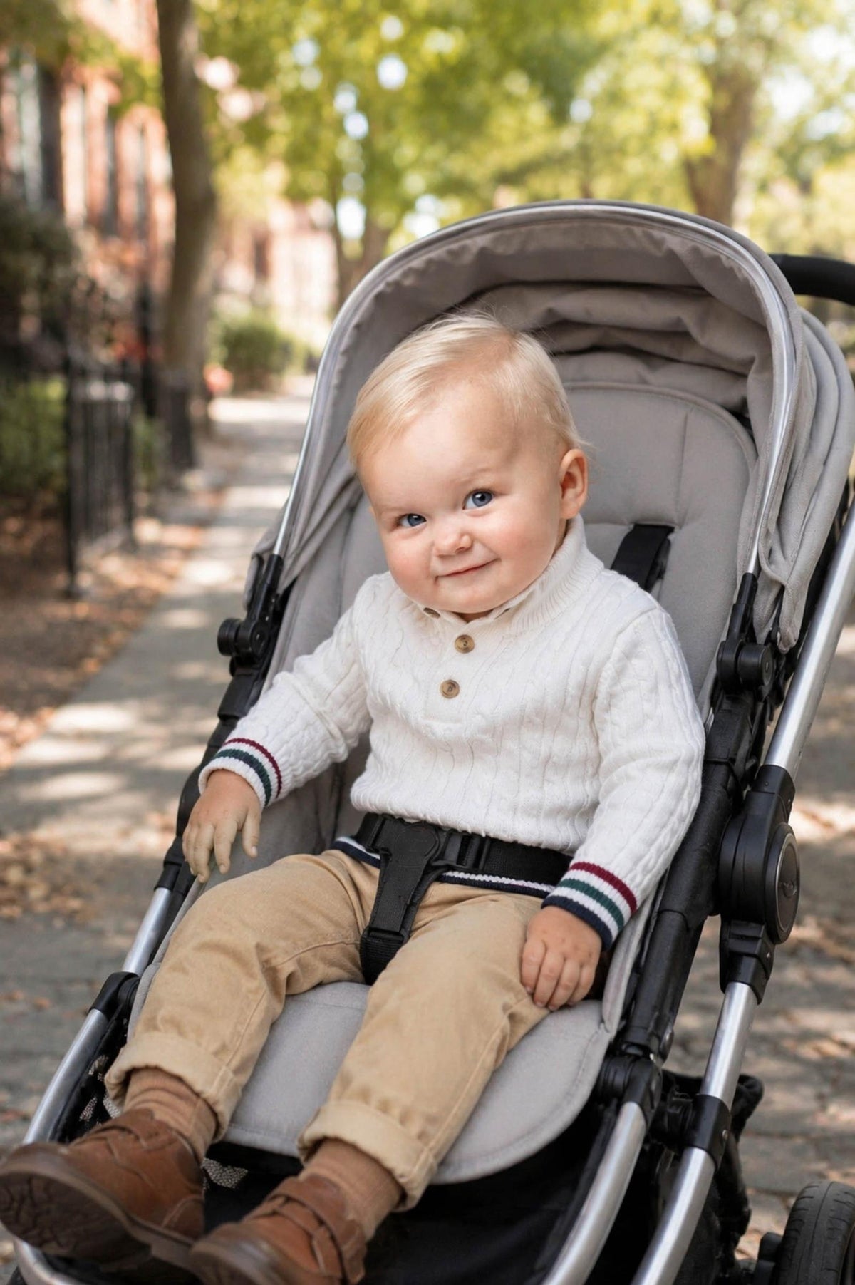 A happy toddler sits in a stroller on a tree-lined street, wearing a white organic cotton mock neck cable sweater, beige pants, and brown boots, enjoying a sunny day outdoors.