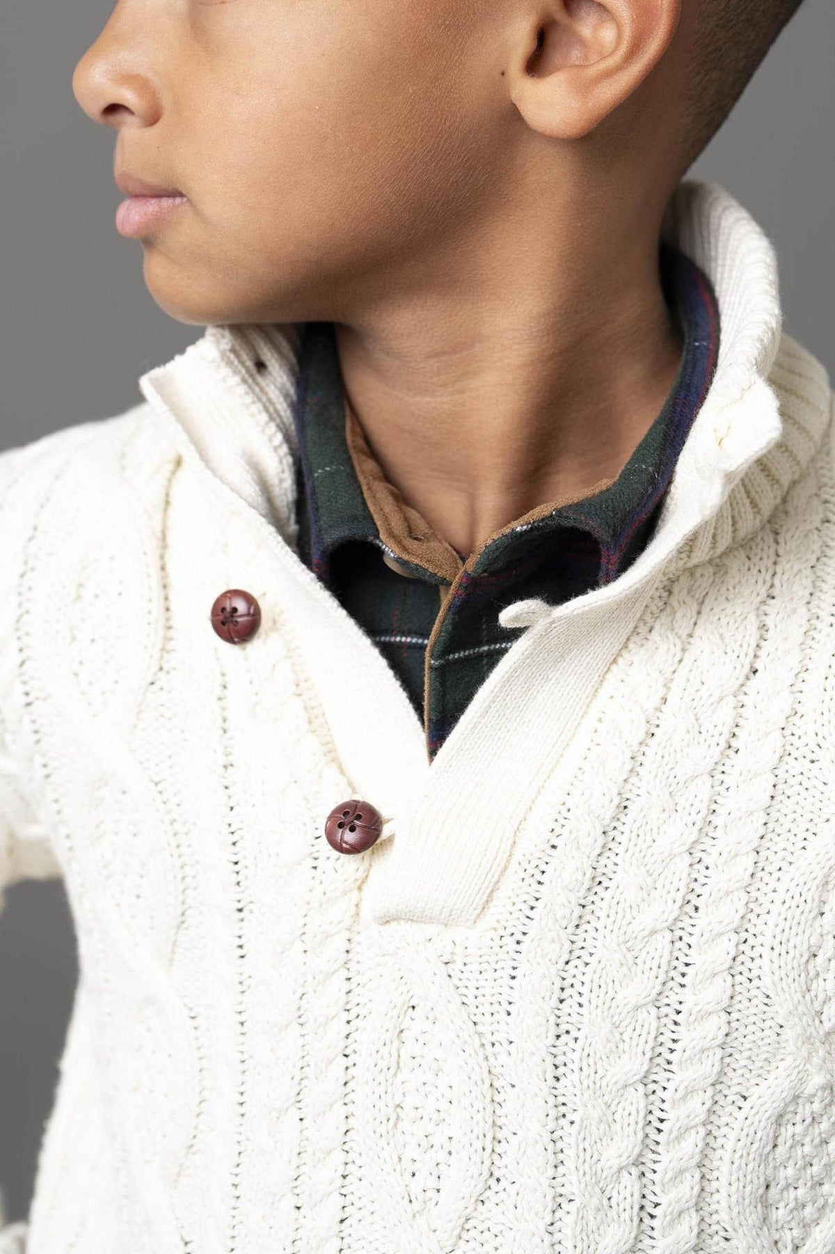 A young boy is wearing a white cable knit mock neck sweater featuring brown buttons. The sweater is made from organic cotton, highlighting a stylish and comfortable look.