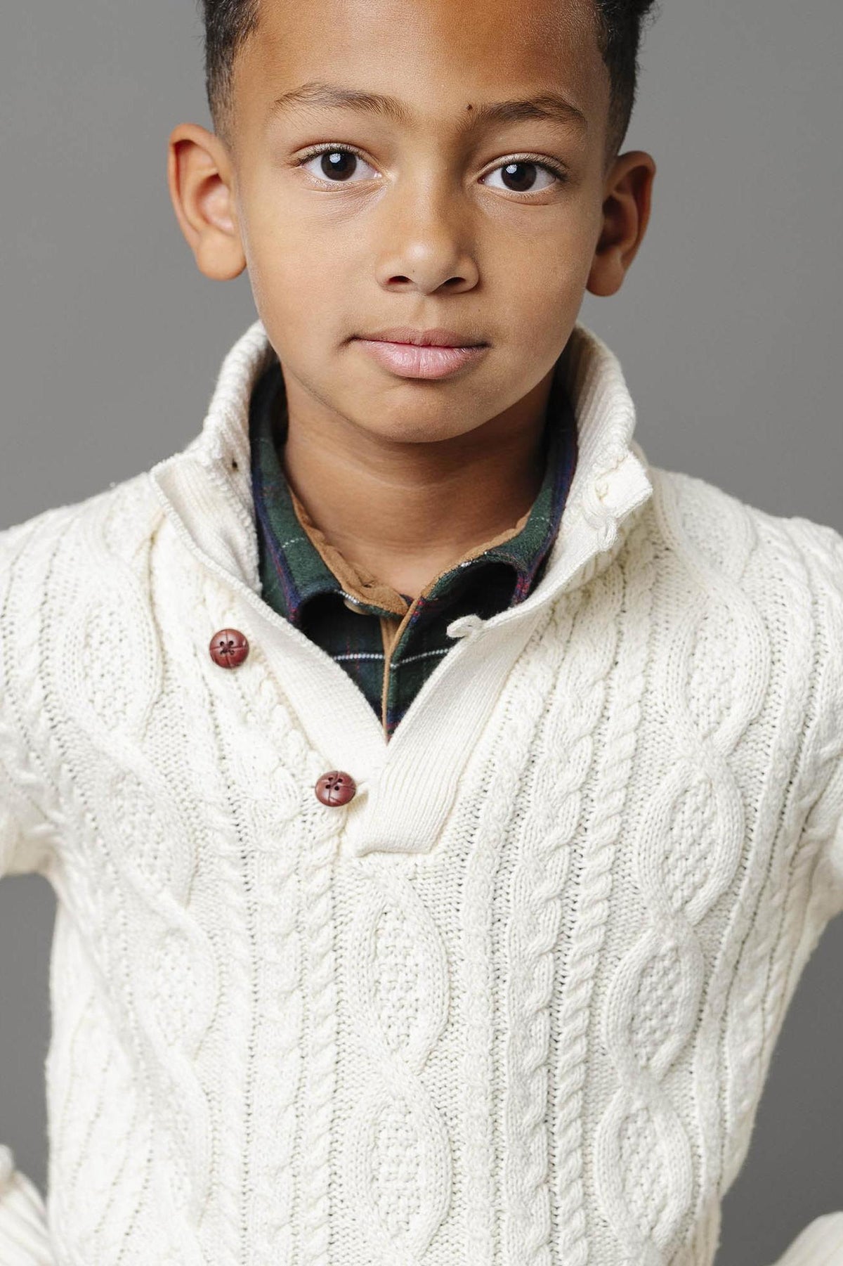 A young boy models a white organic cotton cable knit mock neck sweater with buttons, showcasing its cozy texture and stylish design against a neutral gray background.