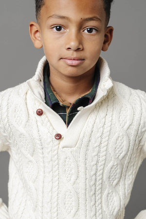 A young boy models a white organic cotton cable knit mock neck sweater with buttons, showcasing its cozy texture and stylish design against a neutral gray background.