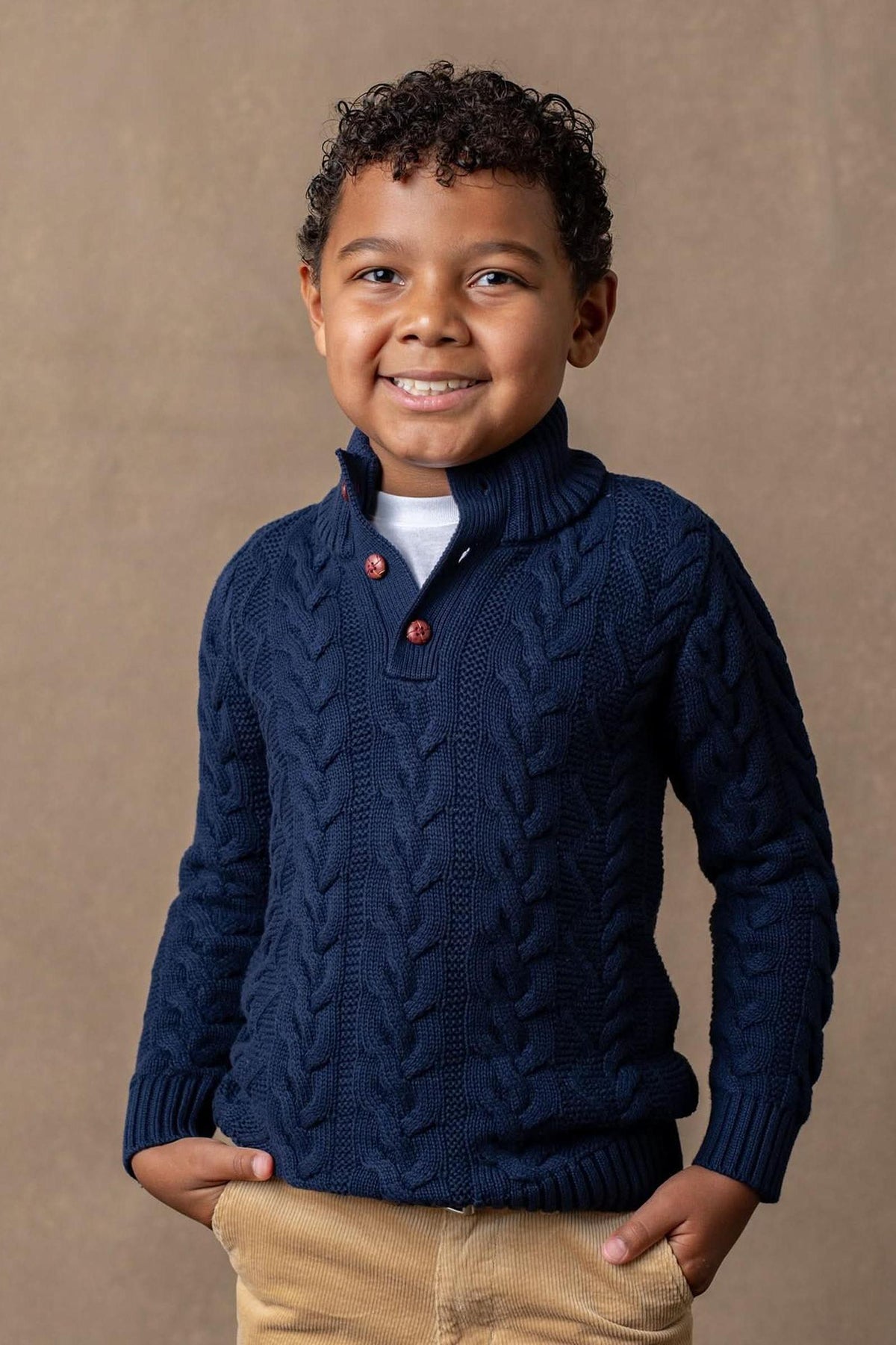 A young boy smiles while wearing a navy cable knit sweater with a mock neck and faux leather buttons.