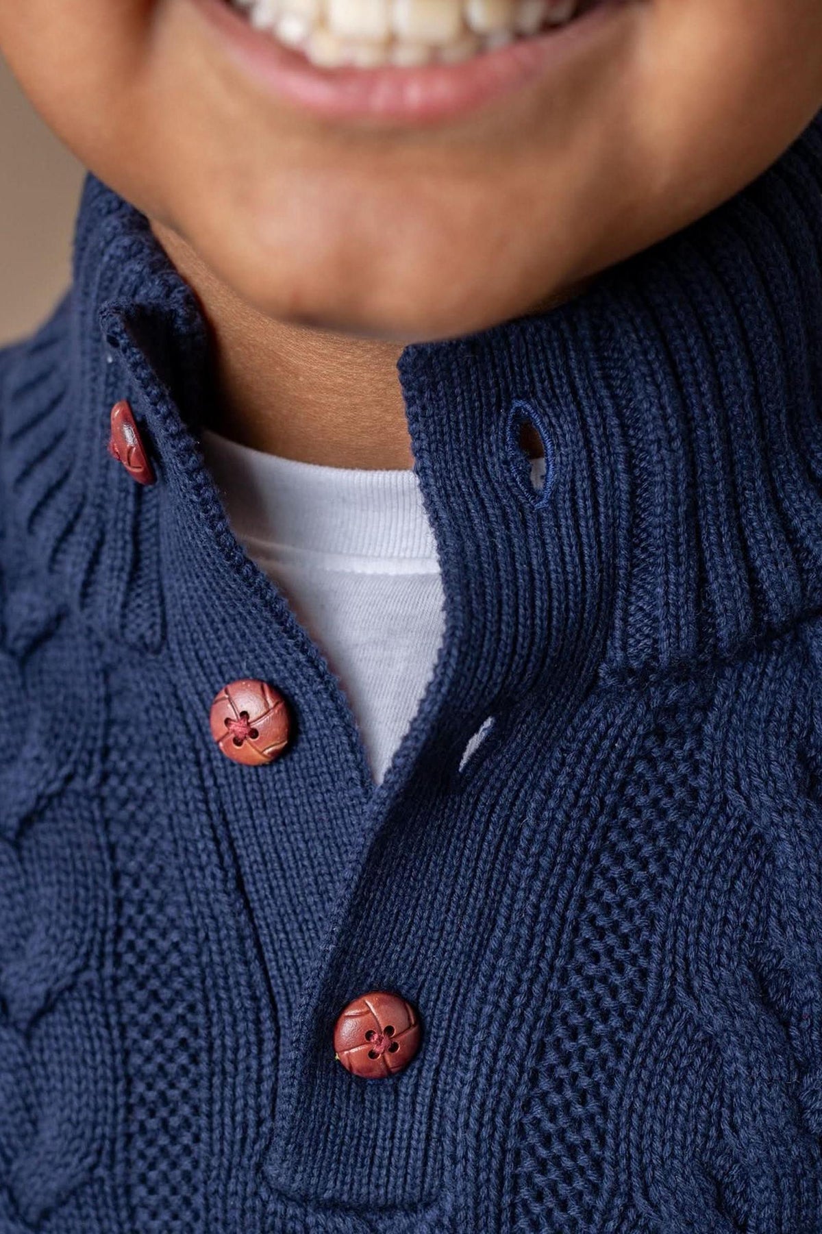 A close-up of a smiling child wearing a navy cable knit sweater with a mock neck and red faux leather.