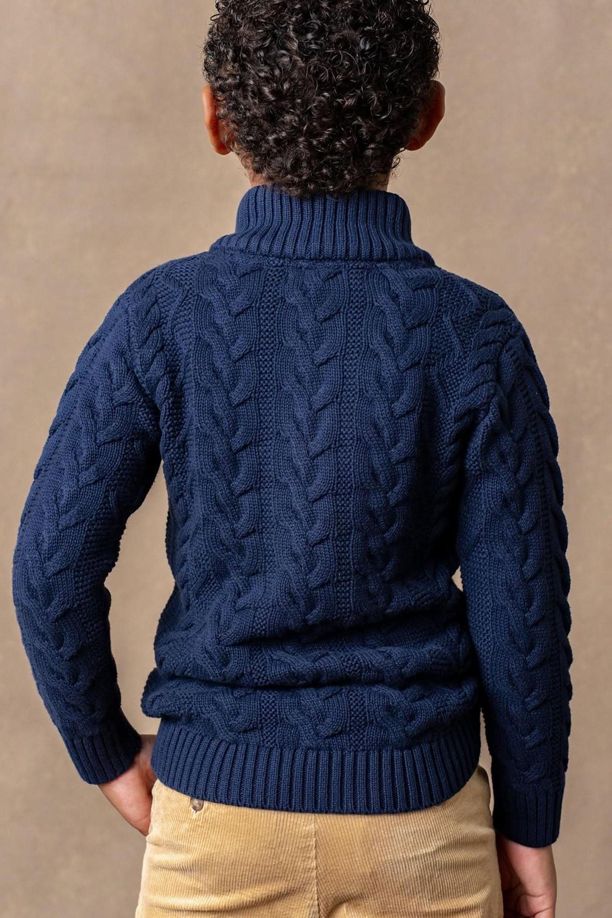 A boys navy mock neck cable sweater features plush cable knit detailing, paired with beige corduroy pants against a neutral.