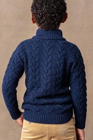 A boys navy mock neck cable sweater features plush cable knit detailing, paired with beige corduroy pants against a neutral.