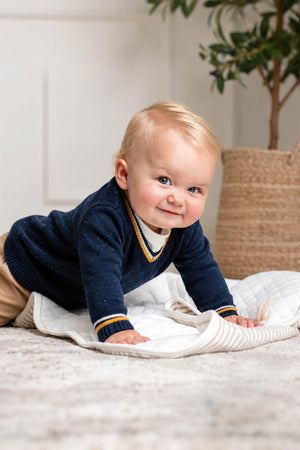 A baby boy with blonde hair is crawling on a soft blanket, wearing a navy organic cotton v-neck sweater with a striped neckline.