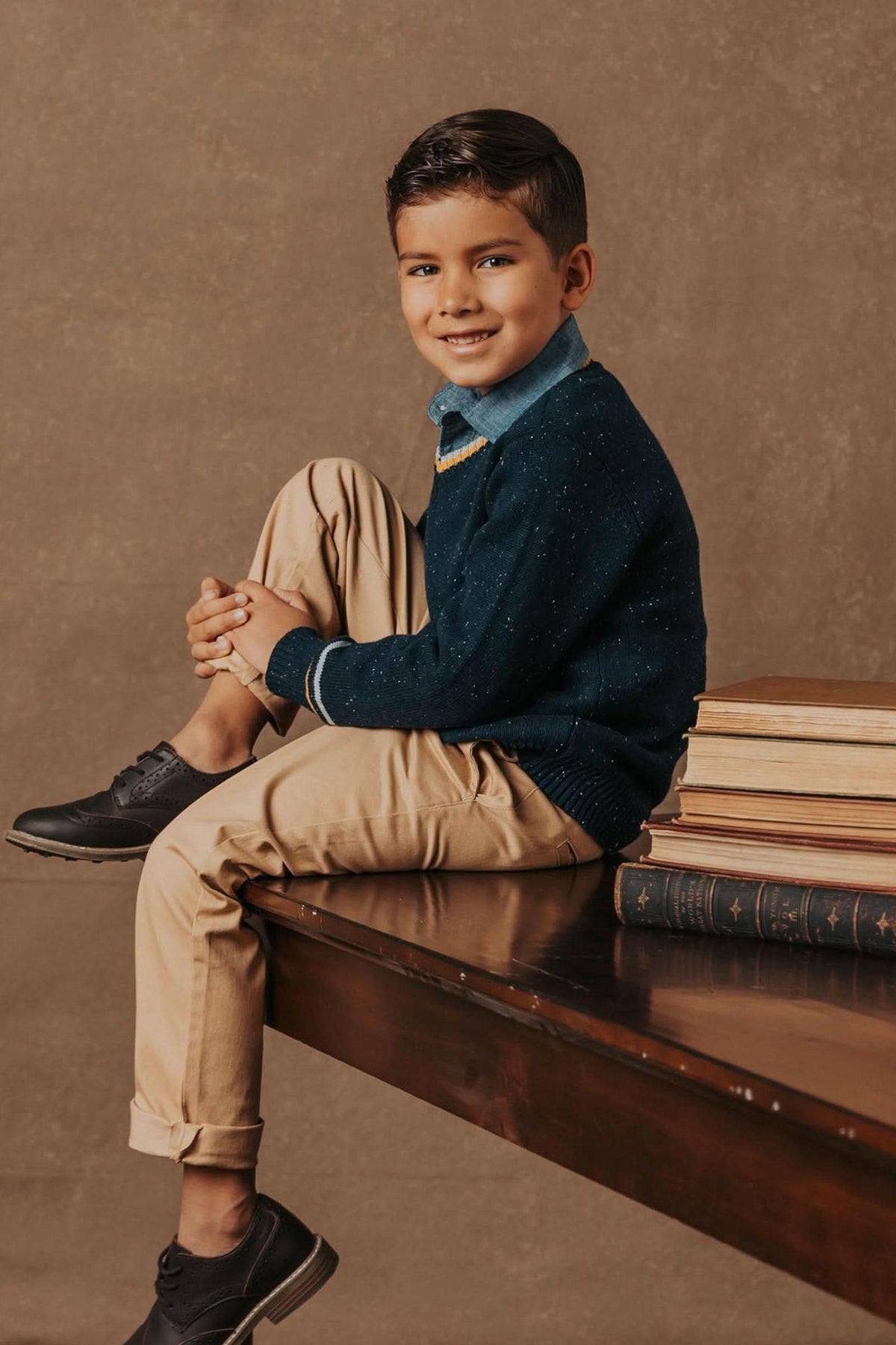 A young boy sits on a wooden table wearing a navy sweater and beige pants, with books stacked beside him.