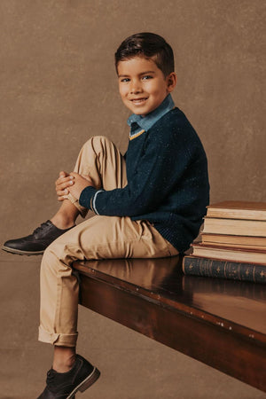 A young boy sits on a wooden table wearing a navy sweater and beige pants, with books stacked beside him.