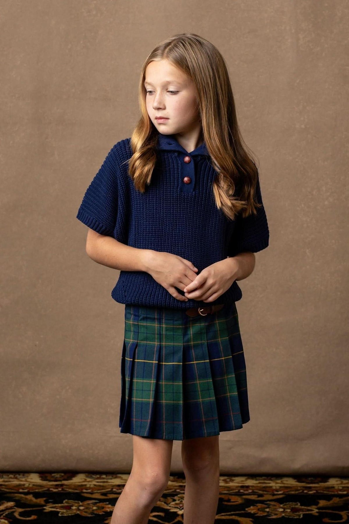 A girl stands wearing a navy knitted shirt and a navy plaid pleated skirt, set against a textured brown background.