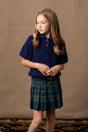 A girl stands wearing a navy knitted shirt and a navy plaid pleated skirt, set against a textured brown background.