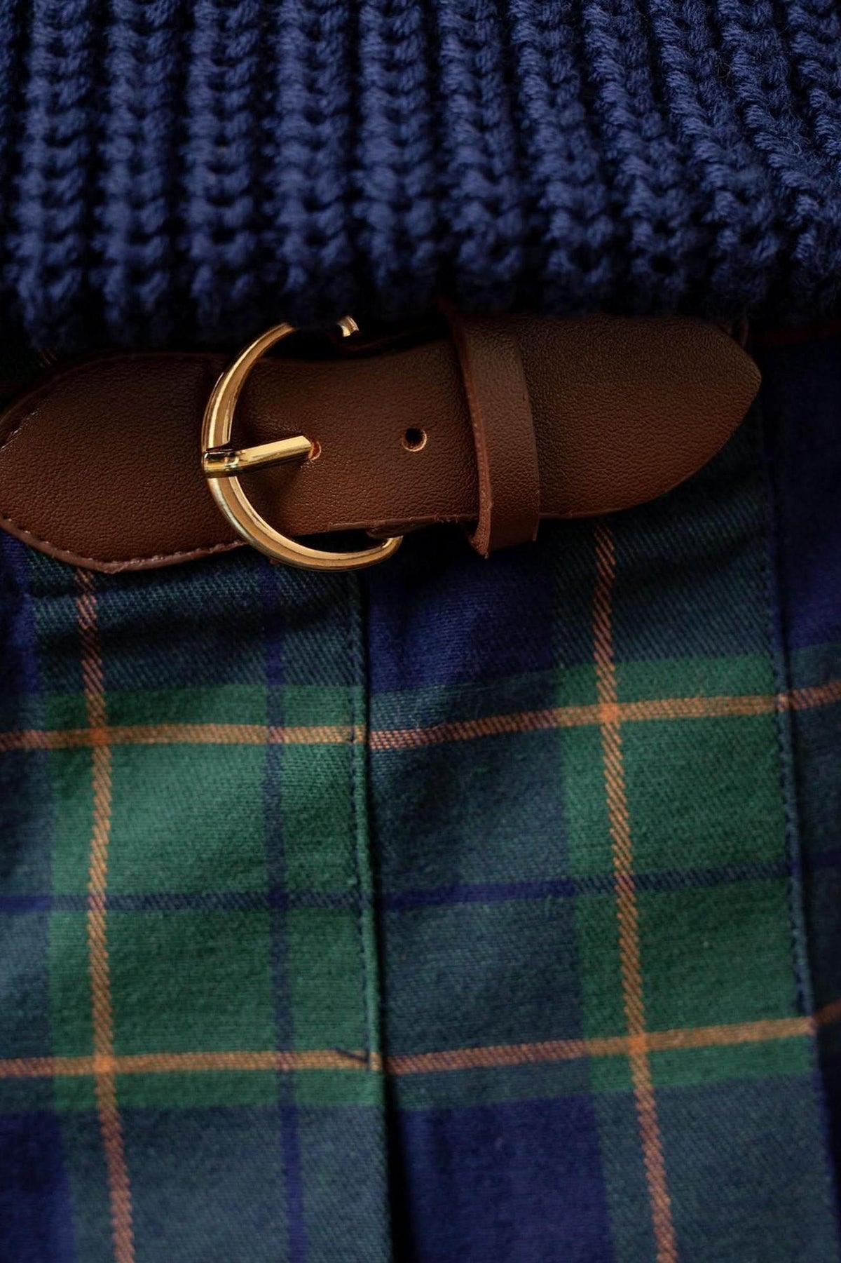 Close-up of a navy plaid skirt featuring a brown faux leather buckle and gold detailing, showcasing its pleated design.
