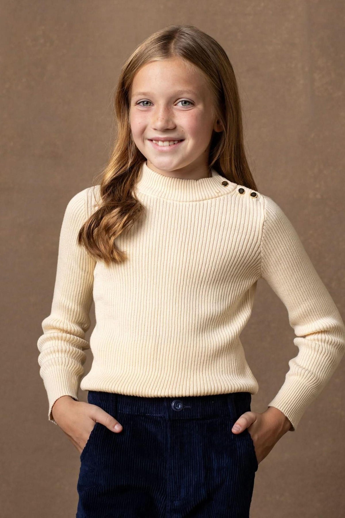 A young girl smiles while wearing a soft ivory mock neck sweater with ribbed texture and gold button details.