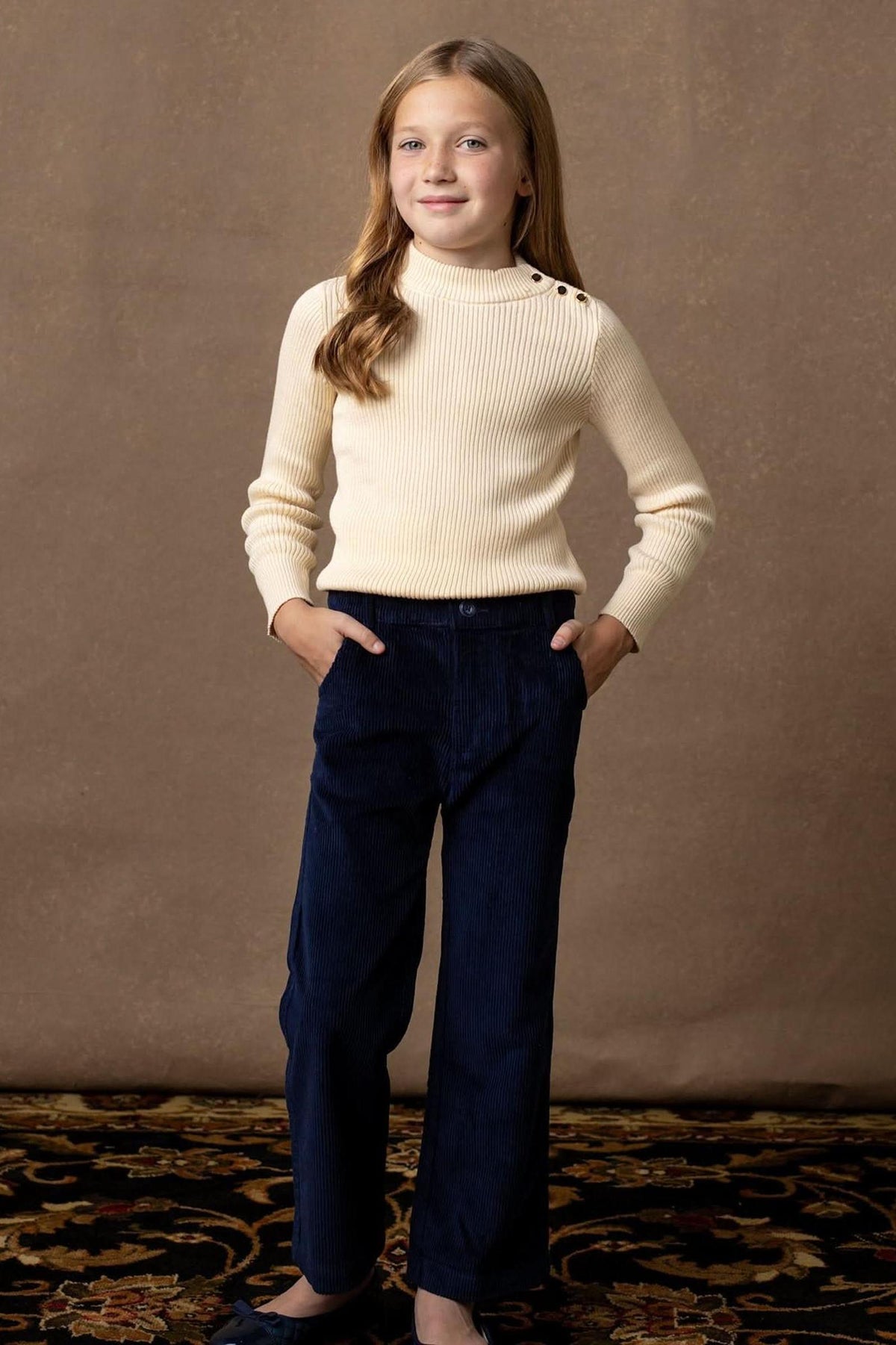 A young girl poses confidently wearing a soft ivory mock neck sweater with rib knit trim and gold button details, paired with dark navy corduroy pants against a neutral backdrop.