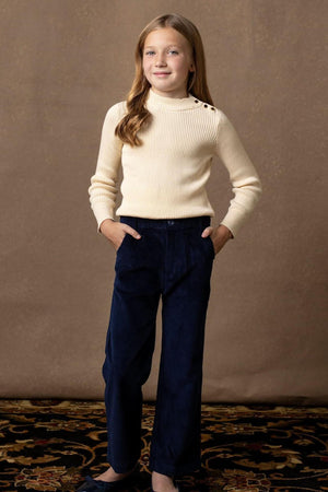 A young girl poses confidently wearing a soft ivory mock neck sweater with rib knit trim and gold button details, paired with dark navy corduroy pants against a neutral backdrop.
