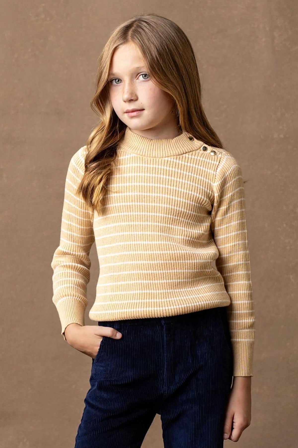 A young girl models a striped mock neck sweater in soft oatmeal and cream hues, featuring button details on the shoulder.