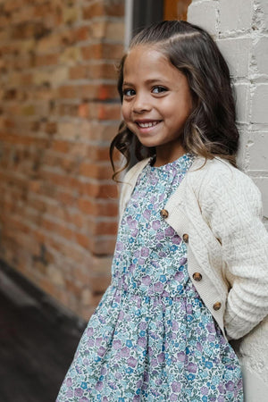 A smiling girl wears a cream cable-knit cardigan over a floral dress, standing against a brick wall.