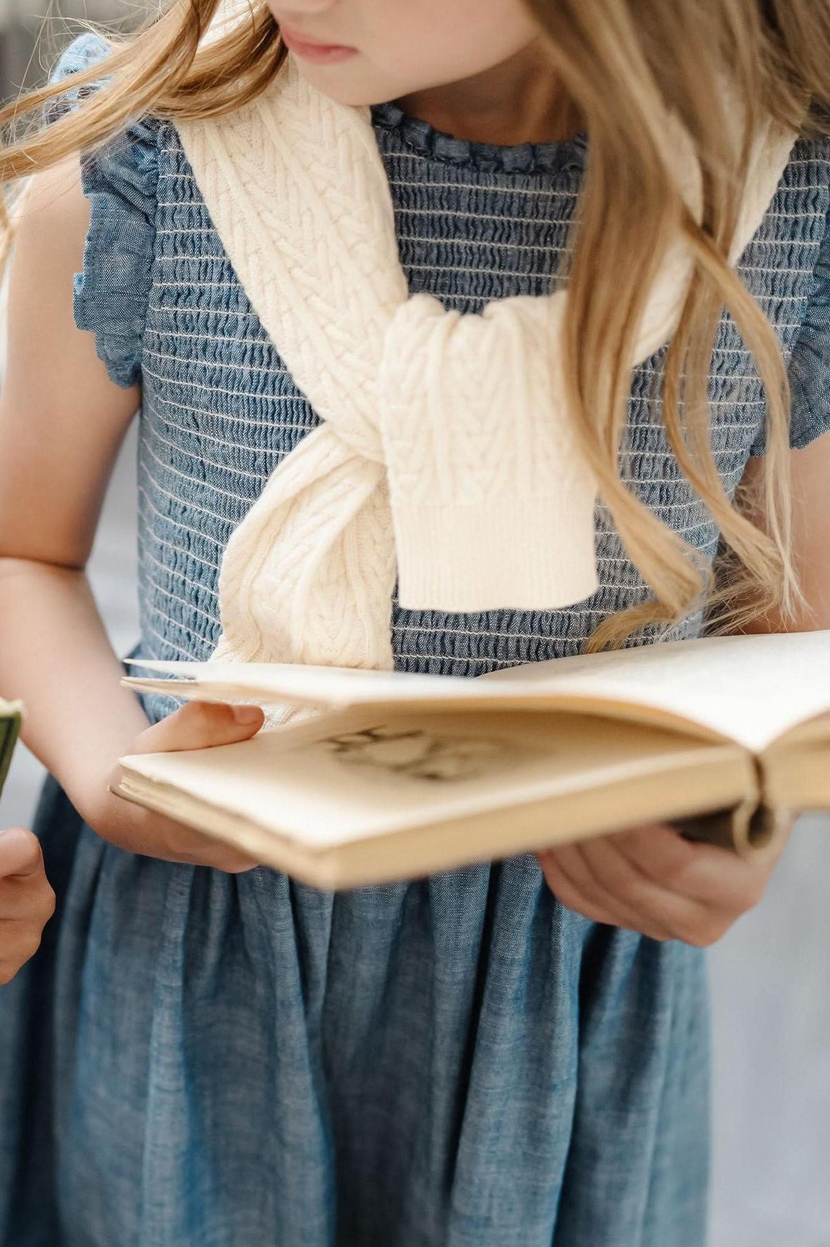 A girl wearing a blue dress holds an open book, with a cream-colored cable-knit cardigan draped around her shoulders.