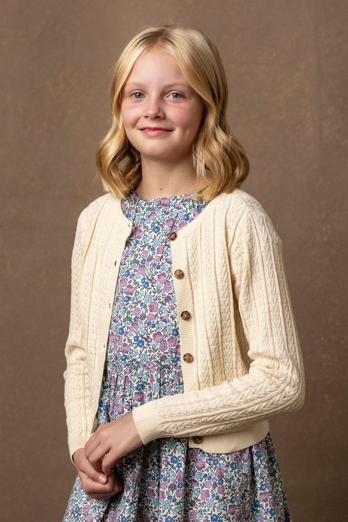 A young girl smiles while wearing an ivory cable-knit cardigan over a floral dress, set against a neutral background.
