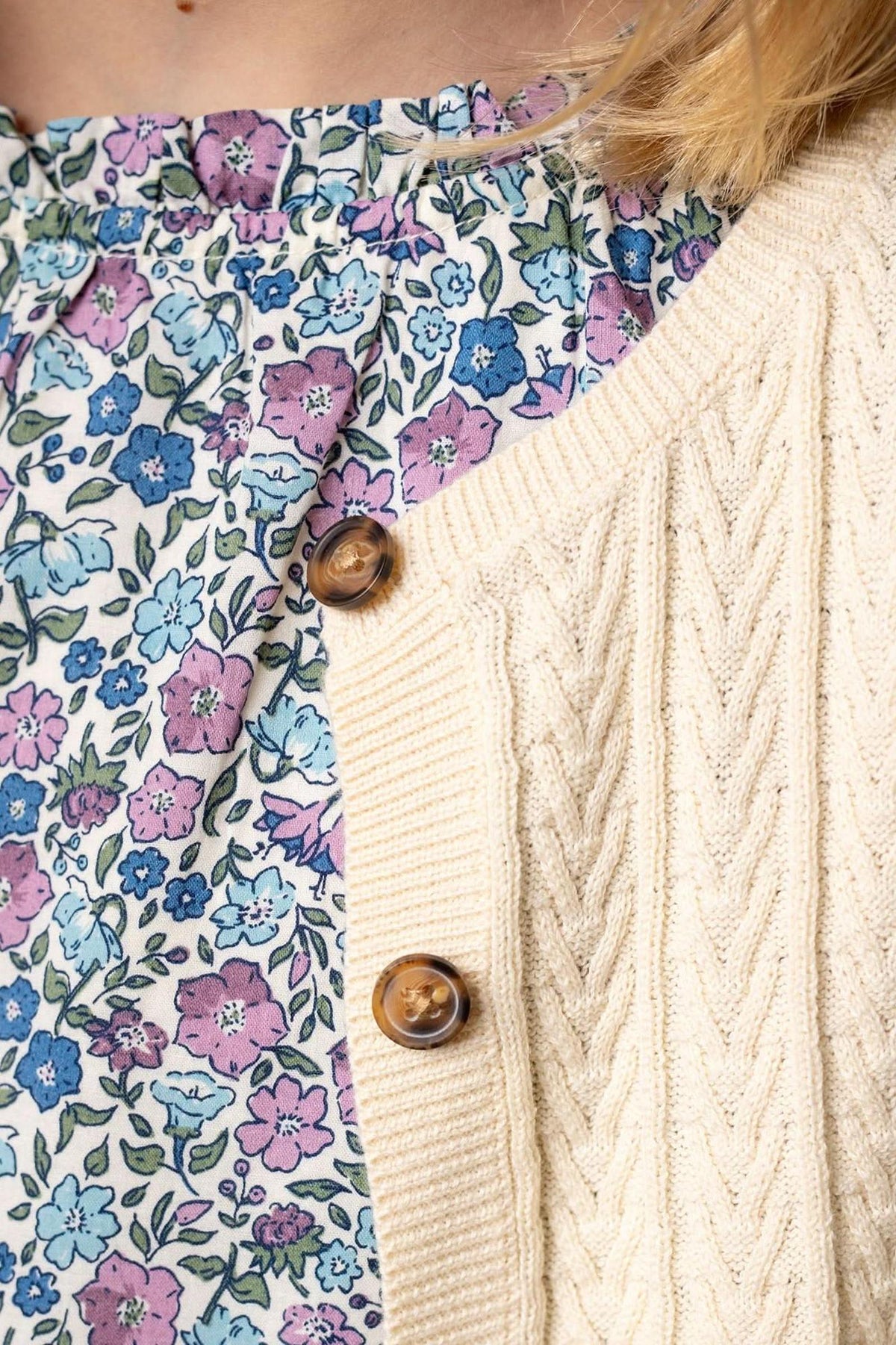 Close-up of an ivory cable knit cardigan with faux horn buttons layered over a floral patterned top in pastel colors.