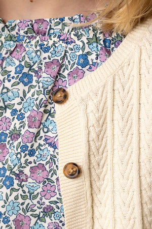 Close-up of an ivory cable knit cardigan with faux horn buttons layered over a floral patterned top in pastel colors.