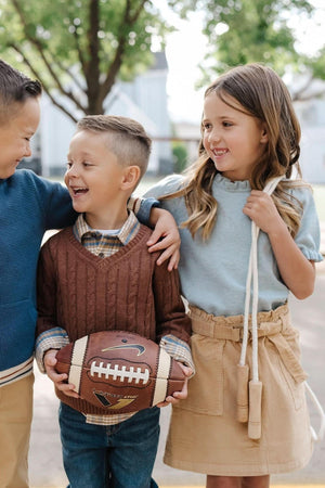 Three children are smiling and interacting outdoors, with one boy holding a brown football. The girl on the right wears a khaki cinch waist skirt, showcasing a casual, playful style.