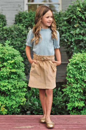 A young girl stands outdoors, wearing a light blue top with puffed sleeves and a khaki corduroy skirt featuring a cinched waist and side pockets, complemented by shiny gold flats.