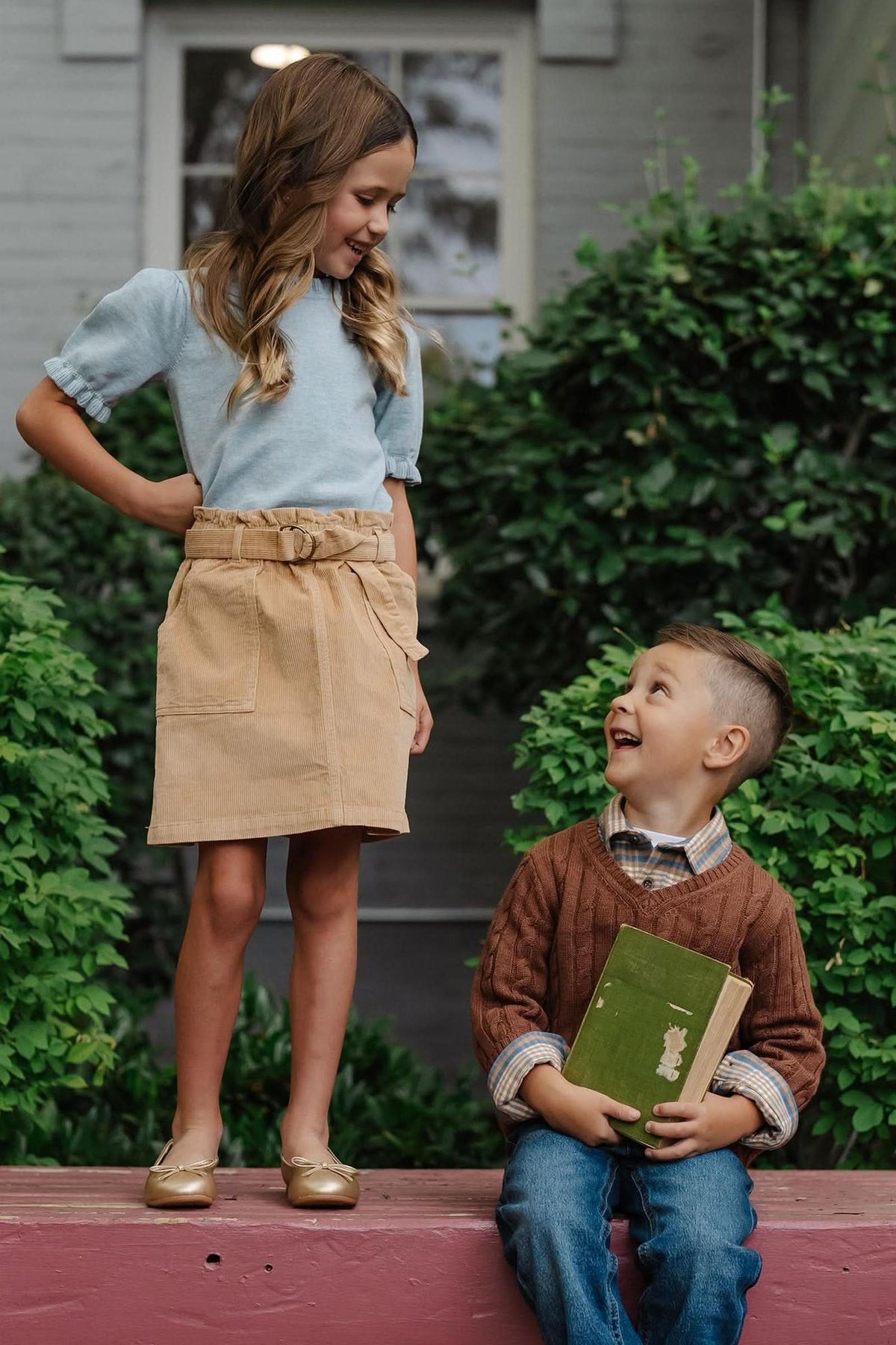 A girl wearing a khaki corduroy skirt with a cinched waist stands on a ledge, smiling at a boy holding a book.