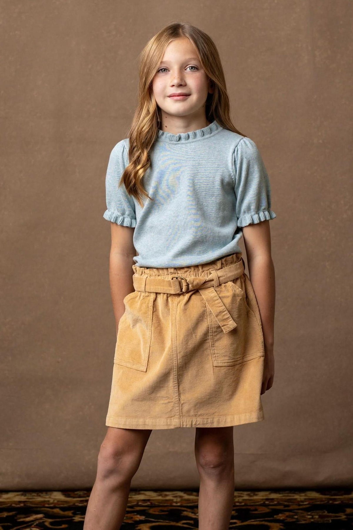 A young girl stands against a neutral backdrop, wearing a light blue ruffled short-sleeve top and a khaki organic cotton corduroy skirt with a cinched waist and side pockets.