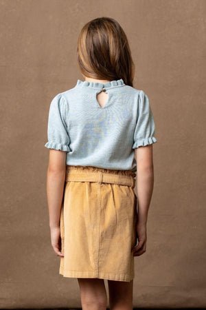 A girl stands with her back to the camera, wearing a light blue top with puffed sleeves and a keyhole detail.