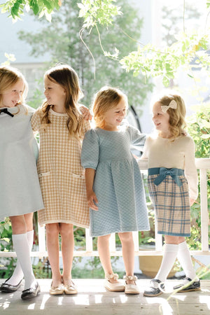 Four young girls stand together wearing stylish dresses in soft colors, enjoying a sunny outdoor setting with greenery.