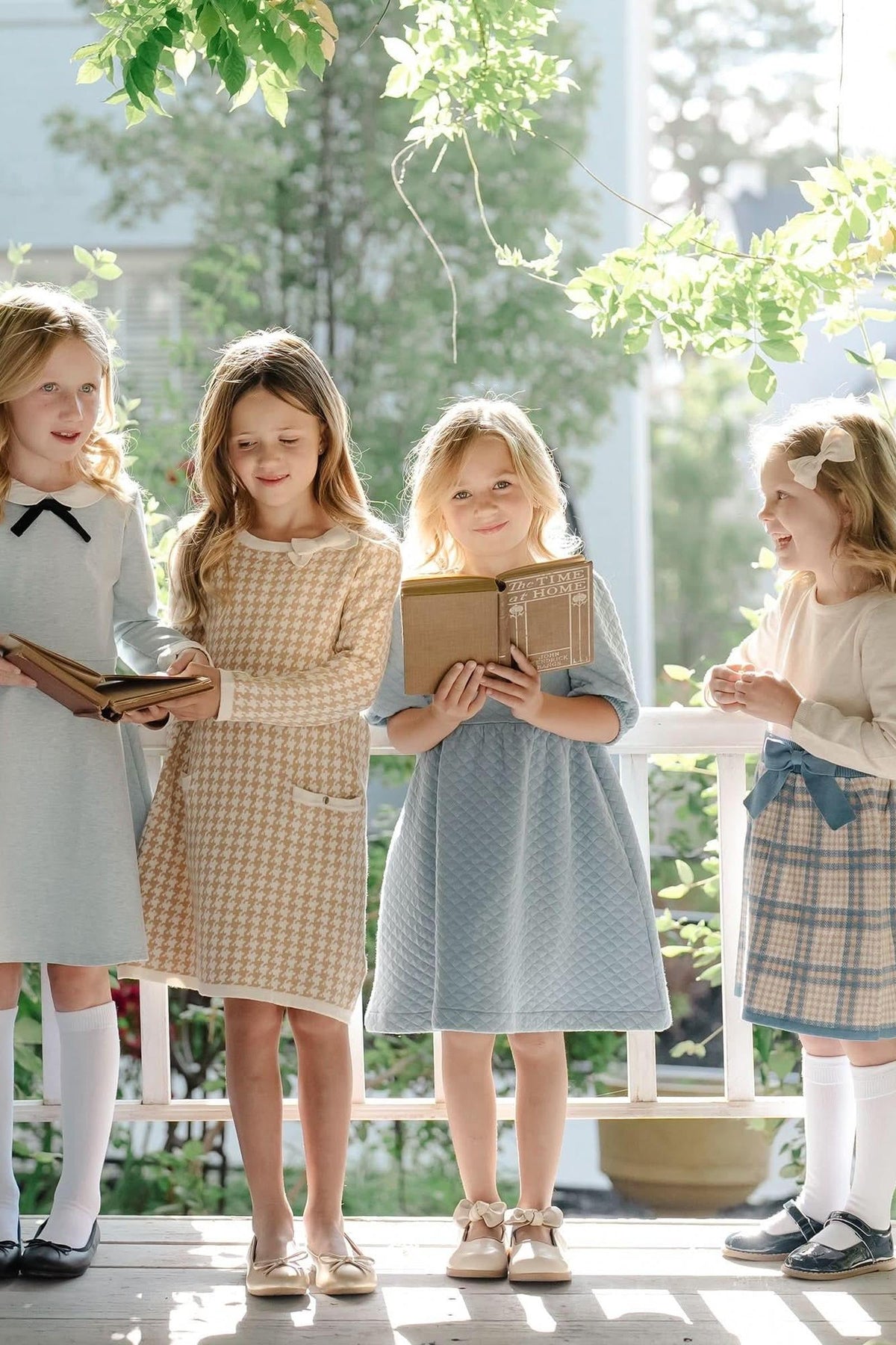 Four girls in stylish dresses stand on a porch, holding books. The setting is bright with greenery in the background.