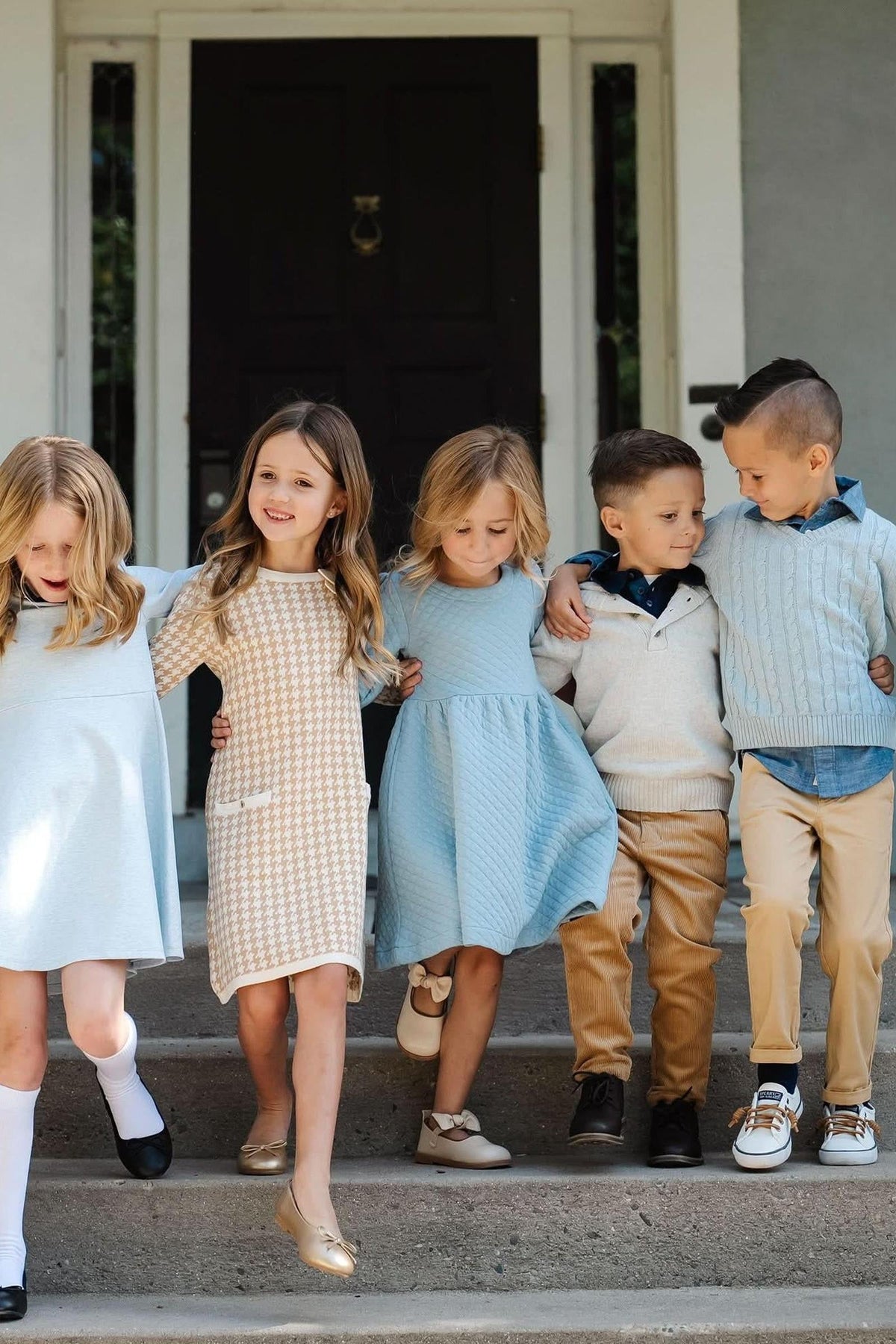 A group of children wearing a variety of stylish dresses and outfits, standing on steps in a cheerful setting.