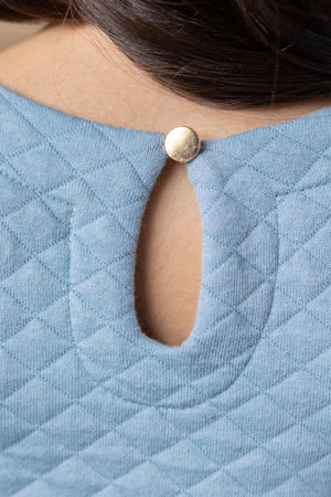 Close-up of a dusty blue quilted fabric with a gold button keyhole closure at the back, showcasing soft, organic cotton.