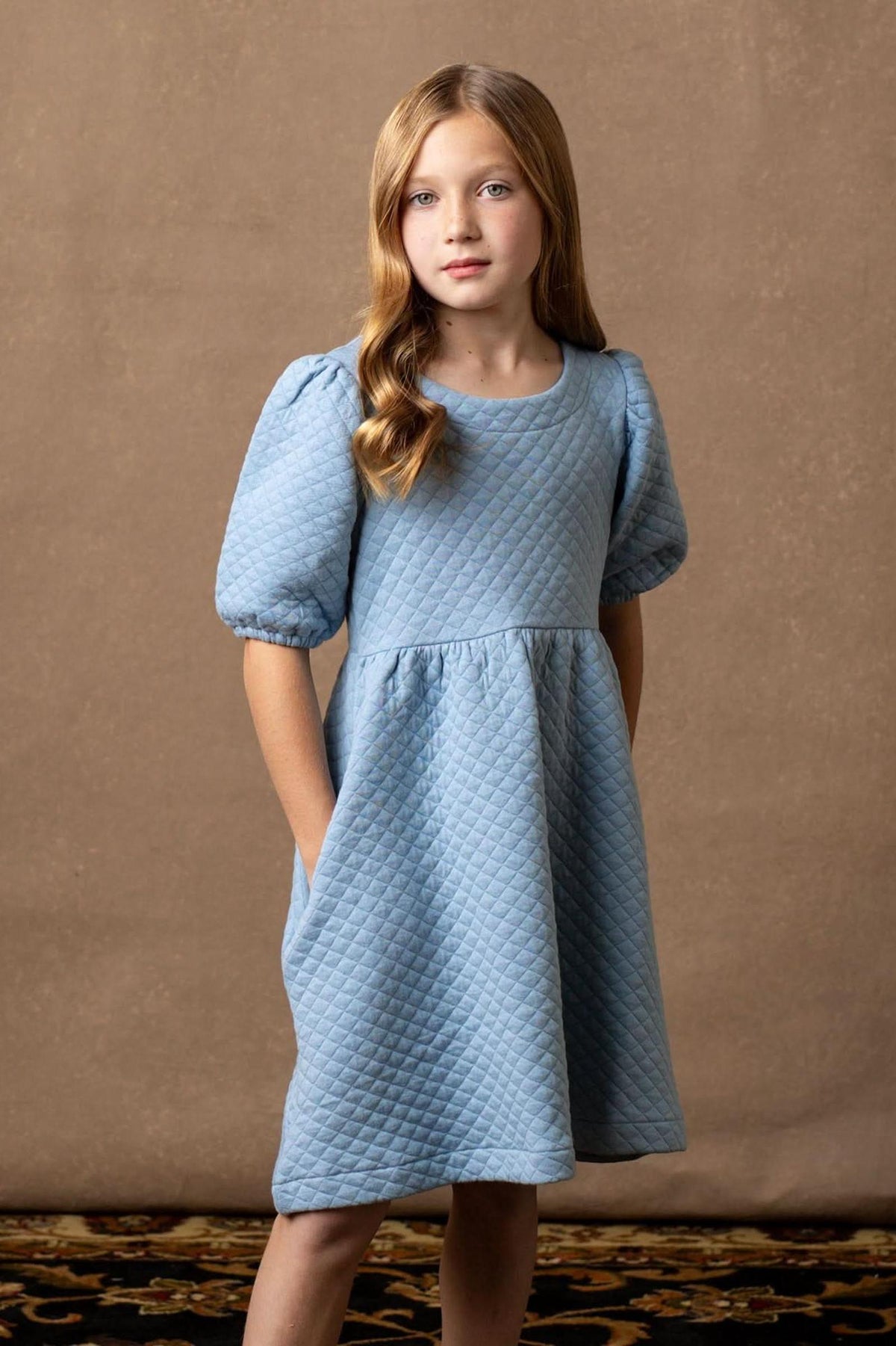A young girl poses in a dusty blue quilted dress with puff sleeves, showcasing a comfortable and stylish silhouette.