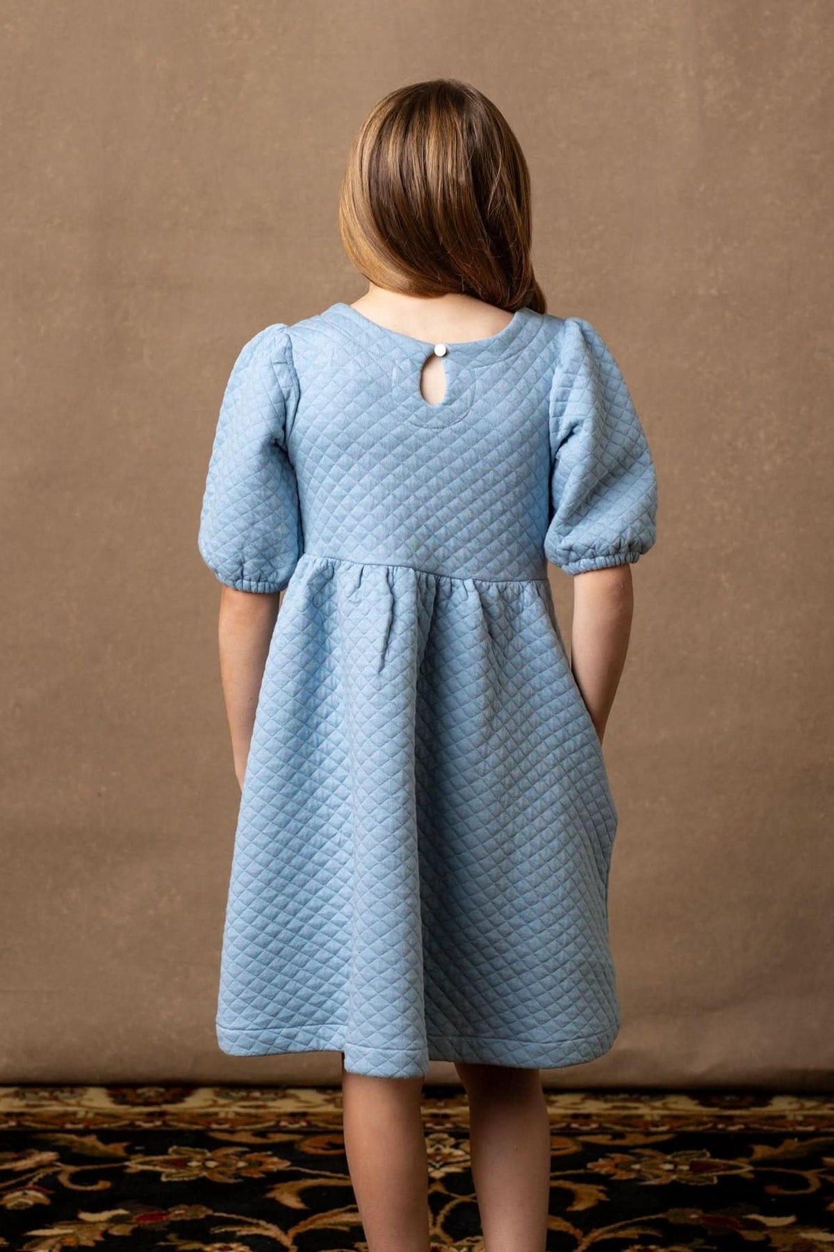 A girl wearing a dusty blue quilted dress with puff sleeves stands against a neutral background, showcasing a keyhole closure.