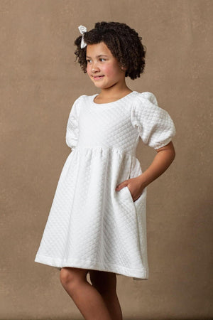 A young girl models a soft white quilted dress with puff sleeves and pockets, set against a neutral brown background.