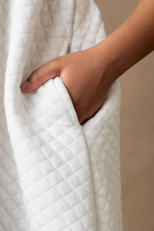 A childs hand is tucked into the pocket of a soft white quilted fabric, showcasing the dresss textured material.