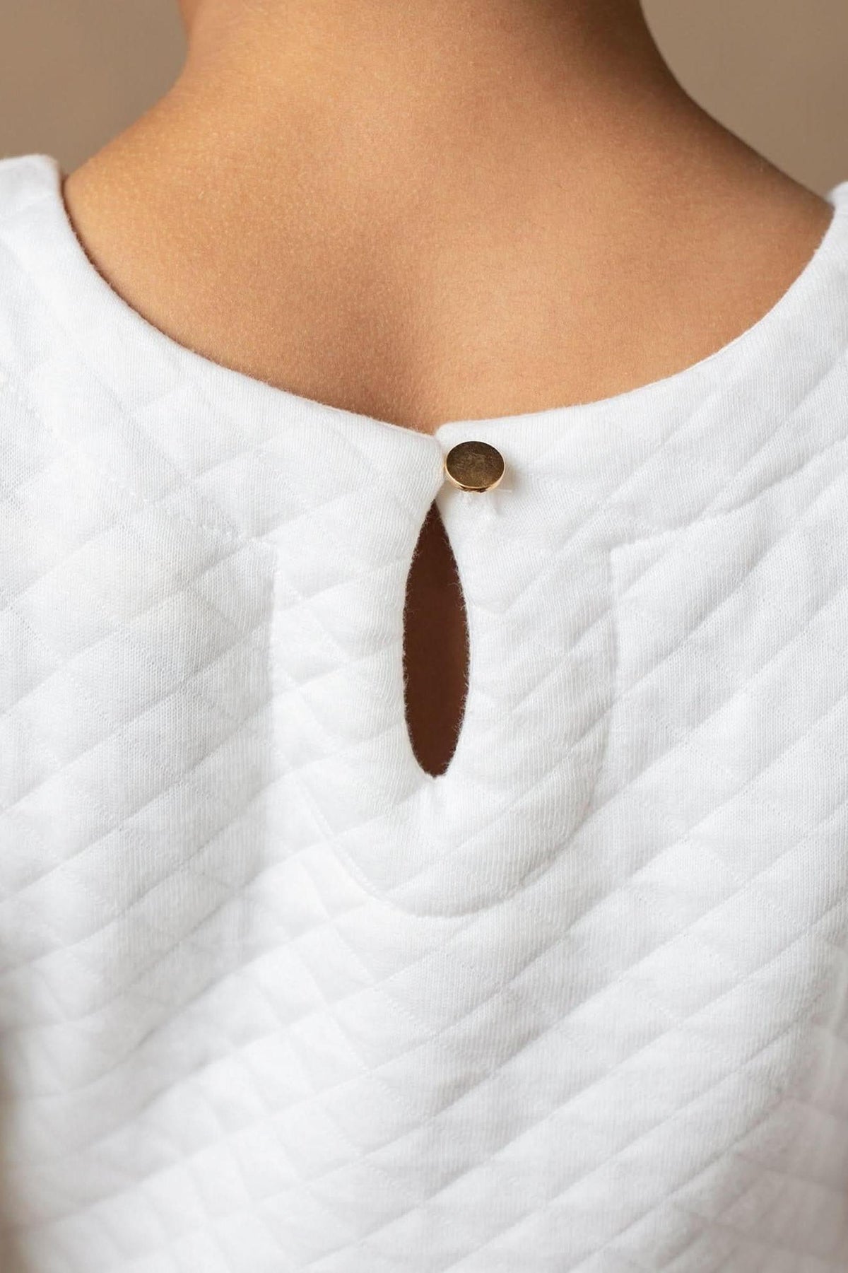 Close-up of a soft white quilted dress showing a gold button closure and keyhole detail at the back neckline.