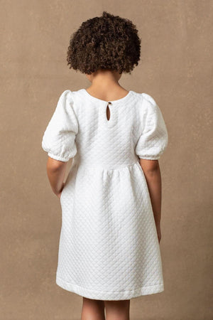 A young child is wearing a soft white quilted dress with puff sleeves, featuring a keyhole closure at the back.