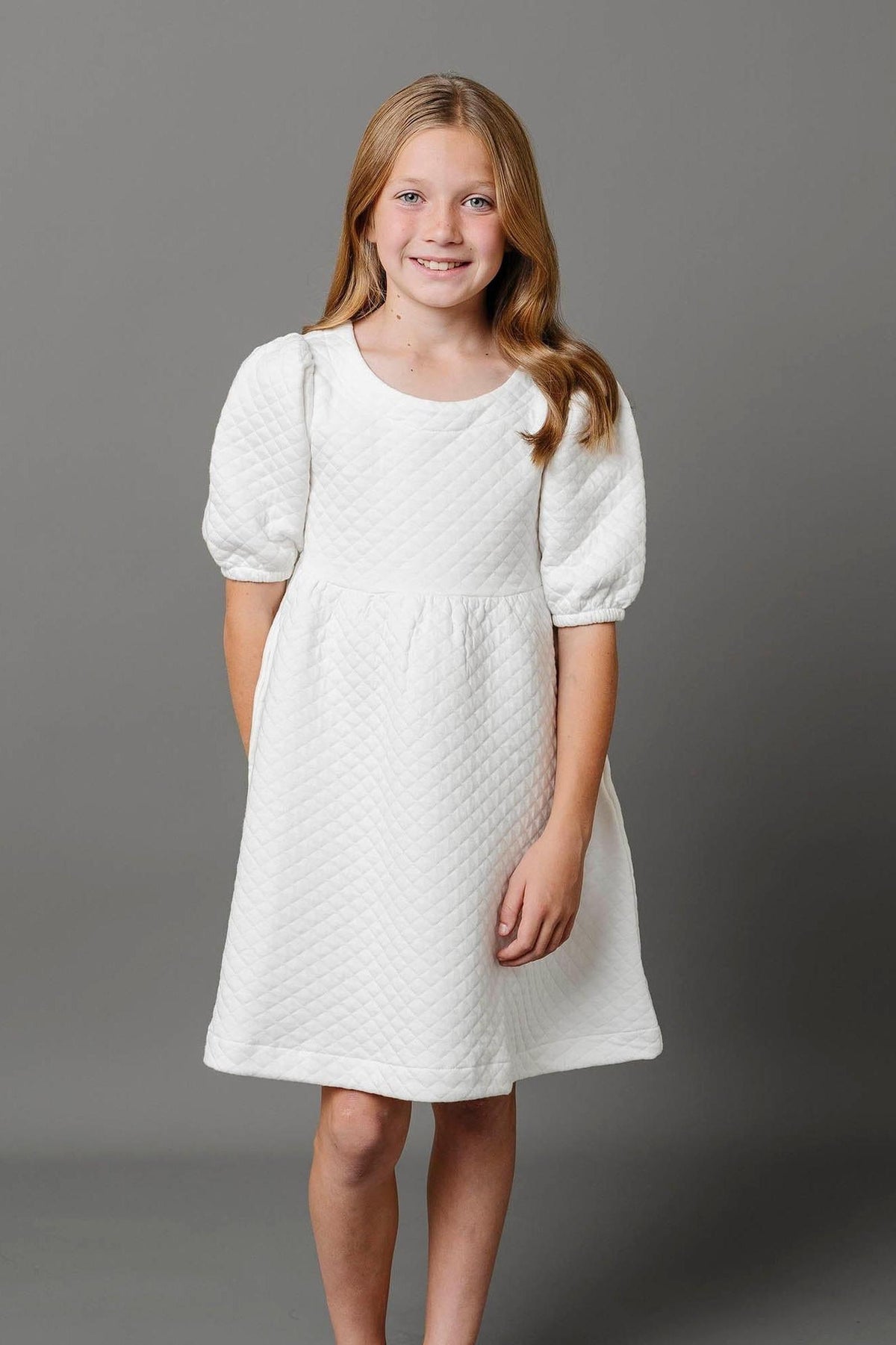 A girl smiles while wearing a soft white quilted dress with puff sleeves, standing against a gray background.