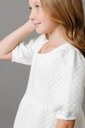 A young girl models a soft white quilted dress with puff sleeves, showcasing its textured fabric and elegant silhouette.