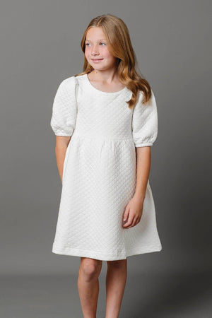 A young girl smiles while wearing a soft white quilted dress with puff sleeves, set against a gray background.