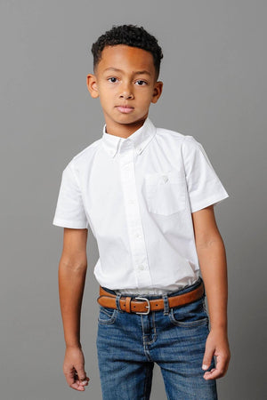 A boy wearing a white short-sleeve button-down shirt and jeans stands against a gray background.